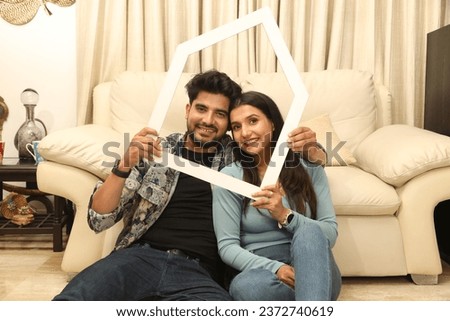 Indian happy young couple holding Papercut home shape frame while sitting on living room and looking in front of the camera Concept - Luxurious lifestyle 
Real Estate in india. Royalty-Free Stock Photo #2372740619