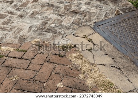 Metal utility cover and multiple brick herringbone patterns with patches of new concrete form a creative copy space background, dead grass in the cracks, horizontal aspect
