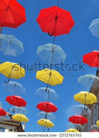 A street decorated with colorful umbrellas during a folk festival. Red, yellow and transparent umbrellas shine above the heads of pedestrians against a background of blue sky. 