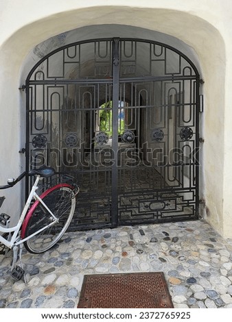 An arched passage through an old house with a ornate wrought-iron gate, through which an inner courtyard with greenery can be seen. A bicycle is parked in front of the passage. Radovljica, Slovenia Royalty-Free Stock Photo #2372765925