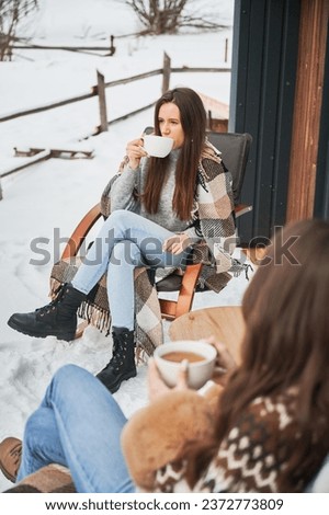 Young women enjoying winter weekends on terrace of contemporary barn house in the mountains. Two girls sitting on chairs and drinking hot tea.