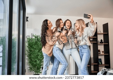 Young women enjoying winter weekends inside contemporary barn house. Four girls having fun, taking selfies on phone and drinking wine.