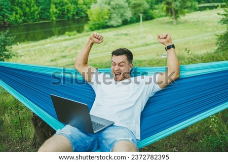 A young man with a laptop sits in a hammock in nature and works remotely. Work in nature during vacation.