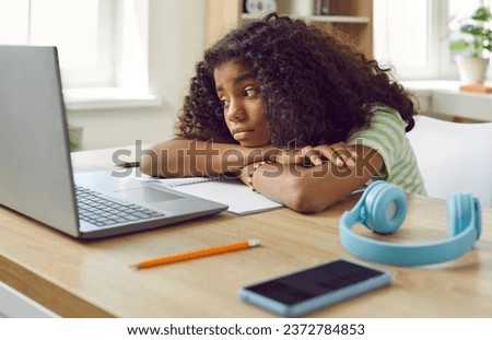 Student gets bored during online lesson. Elementary school child thinking about something else during remote class. Tired, sad diverse Afro American girl sitting at her table with laptop and notebook
