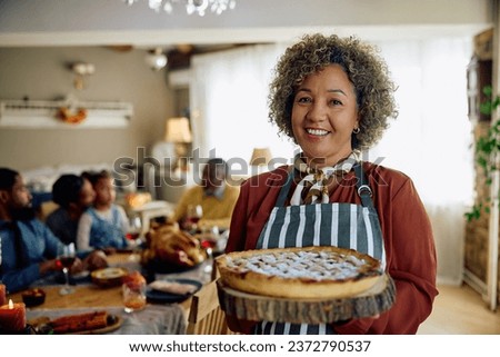 Happy mature woman holding baked Thanksgiving pie and looking at camera. Her family is in the background.