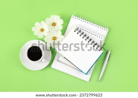 Composition with opened blank notepad and pen, cup of black coffee, flowers of a white cosmos on a green background. Festive office desktop concept. Morning coffee cup. Royalty-Free Stock Photo #2372799623