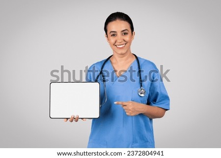 Professional latin woman doctor in blue uniform confidently points towards a digital tablet with blank screen, offering space for customizable content against grey studio background