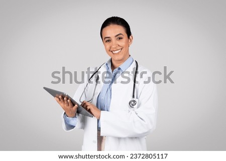 Cheerful latin woman doctor holding clipboard and smiling towards camera, portraying an approachable healthcare professional against grey studio background Royalty-Free Stock Photo #2372805117
