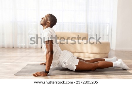 Athletic handsome young black man in sportswear stretching on yoga mat, doing morning workout at home, practicing yoga, copy space, full length candid shot, side view. Healthy lifestyle, sport Royalty-Free Stock Photo #2372805205