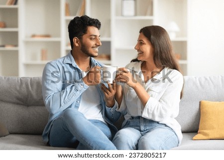 Cheerful young indian couple enjoying leisurely moment on sofa, drinking coffee while having pleasant conversation, creating heartwarming, homey scenario Royalty-Free Stock Photo #2372805217