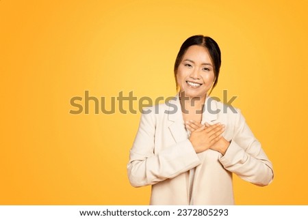Heartfelt moment of smiling Asian millennial woman places hands on chest, eyes gleaming with genuine gratitude and kindness, isolated on orange studio background. People emotions Royalty-Free Stock Photo #2372805293