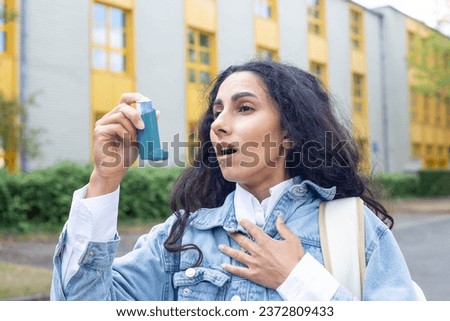 Young Eastern girl has an asthma attack or panic and holds an inhaler in her hand while leaving a school or university building. Inhale vapor from an inhaler - first aid on the street.  Royalty-Free Stock Photo #2372809433