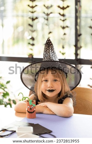 A cute girl in a witch costume makes a paper craft for Halloween. The concept of children's crafts for Halloween.