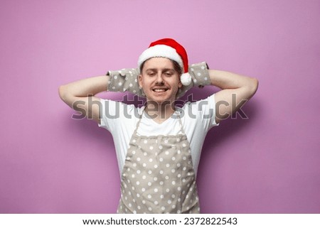 guy chef in santa hat and apron rests and relaxes on pink isolated background, male chef in uniform and baking gloves lies and lazes for the New Year