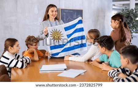 Excited young female teacher explaining culture of country while showing flag of Uruguay to schoolchildren preteens in classroom Royalty-Free Stock Photo #2372825799