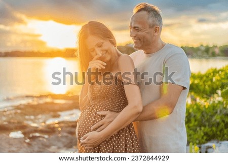A happy, mature couple over 40, enjoying a leisurely walk on the waterfront On the Sunset, their joy evident as they embrace the journey of pregnancy later in life Royalty-Free Stock Photo #2372844929