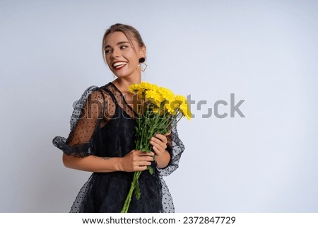 In an elegant black attire and captivating evening makeup, a stunning lady against white wall. Holding a bouquet of sunny yellow flowers, she radiates a festive and cheerful atmosphere