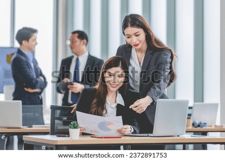 Asian professional successful female businesswoman supervisor mentor in formal suit standing smiling pointing help advising new colleague working with paperwork document at working station in office.