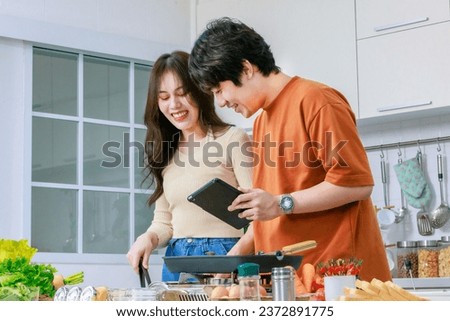Asian young lovely lover couple husband and wife in casual outfit standing smiling holding touchscreen tablet computer learning helping cooking in full decorated modern kitchen with equipment at home.
