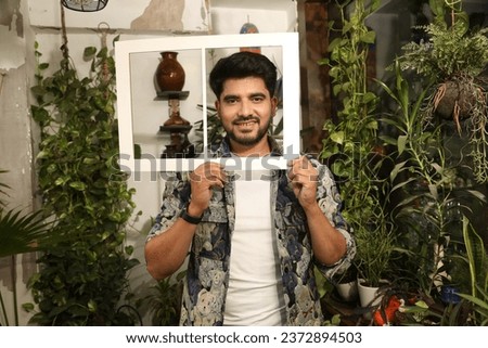Portrait of Young male hold paper photo frame toothy smile look camera isolated greenery balcony at home.
