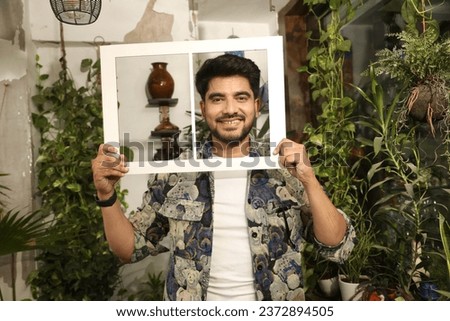 Portrait of Young male hold paper photo frame toothy smile look camera isolated greenery balcony at home.