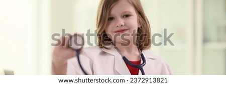 Cute blonde girl holds stethoscope to check patient playing therapist. Little doctor in medical uniform stands in clinic office looking in camera and smiling