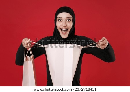 Surprised young arabian muslim woman in hijab black clothes posing isolated on red wall background studio portrait. People religious lifestyle concept. Hold package bag with purchases after shopping