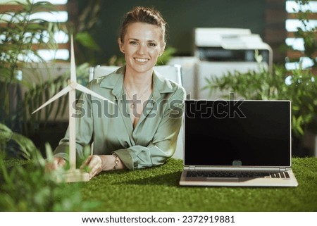 Time to go eco. smiling elegant 40 years old small business owner woman in green blouse in modern green office with windmill showing laptop blank screen Royalty-Free Stock Photo #2372919881