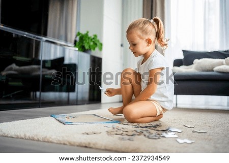 Little blonde girl sits at home on the carpet and collects puzzles. High quality photo
