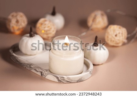 burning candle on marble tray with pumpkins and warm lights on a beige background, autumn atmosphere, cozy home