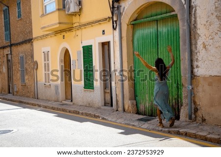 a girl in a green dress of unusual cut walks along the street of a small Spanish town. houses have not been repaired for a long time. the door with green-colored shutters has long been destroyed 