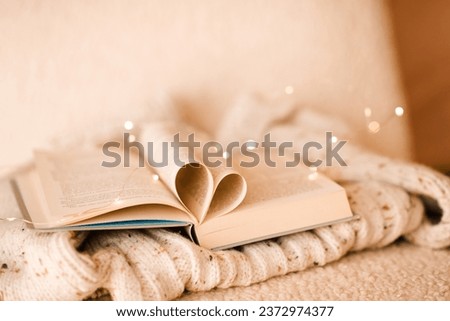 Open paper book with folded pages in heart shape on knitted clothes over Christmas lights in room close up. Winter cozy home atmosphere. 