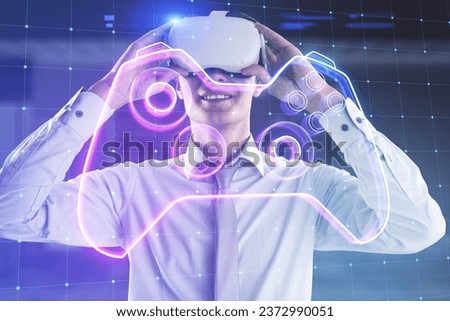Attractive happy guy with VR glasses and creative joystick or gamepad hologram on blurry blue office background. Esport, gaming and fun concept. Double exposure