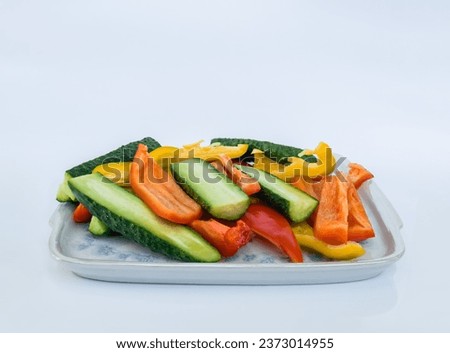 Vegetable slices of yellow and red peppers and cucumbers on a square shaped porcelain dish with rounded corners on a white background