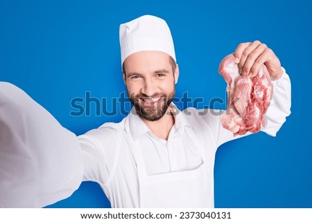 Self portrait of successful joyful butcher with stubble shooting selfie on front camera and holding fresh meat in hand, having video-call, isolated on grey background