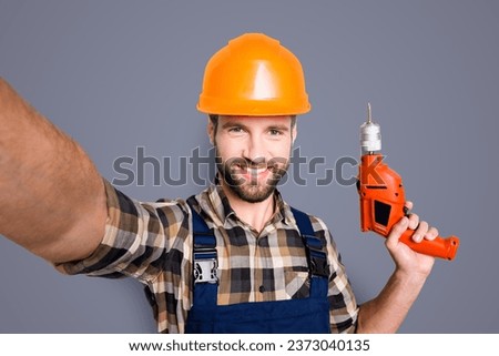 Self portrait of cheerful joyful repairer in protective hard hat shooting selfie on front camera having equipment in hand isolated on grey background. Leisure fun concept
