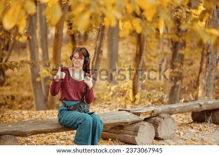 Red-haired teenage girl listens to music on headphones on a bench in an autumn park and shows a thumbs up. Autumn season, yellow foliage.