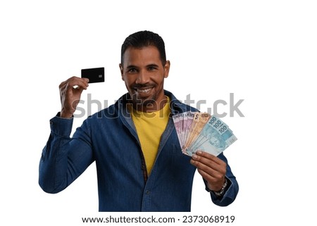 A man is holding Brazilian money and a black credit card, wearing a blue long-sleeved shirt and a yellow T-shirt on a white background.