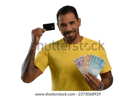 man holds a black credit card and Brazilian money, wearing a yellow shirt on a white background