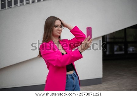 a young business girl in a bright fuchsia jacket stands on the street, looks at the phone, straightens her hair, takes a selfie	

