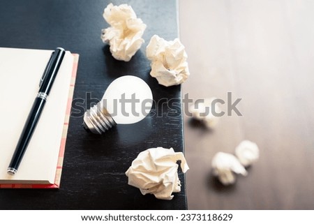 Original idea for writing, closeup light bulb with notebook and crushed paper balls on the desk, and some trash on the ground Royalty-Free Stock Photo #2373118629
