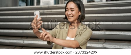 Portrait of asian beautiful girl takes selfie on smartphone. Young korean woman sits on stairs outdoors and makes photos on mobile phone.