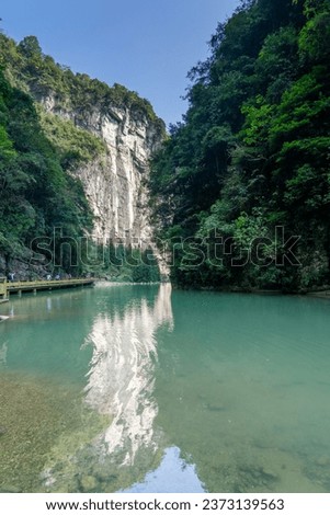 The picture captures the breathtaking beauty of Wulong Karst in China. A turquoise river flows in the foregroundThe right side is adorned with vibrant green trees under a clear blue sky