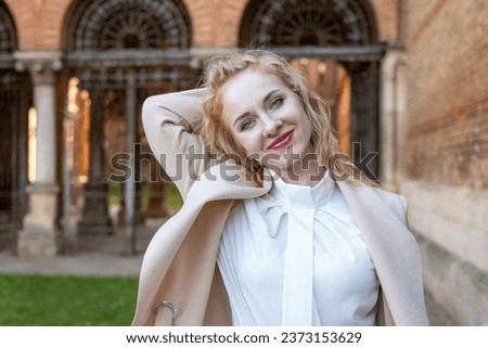 Cute blonde girl in white blouse and coat outside smiling and looking at the camera. Portrait of attractive red-haired girl in spring outdoors