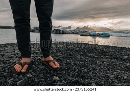 Close-up picture of male feet in slippers standing on the seashore in nature in Iceland. The unknown man is standing on the shore in Iceland in slippers.