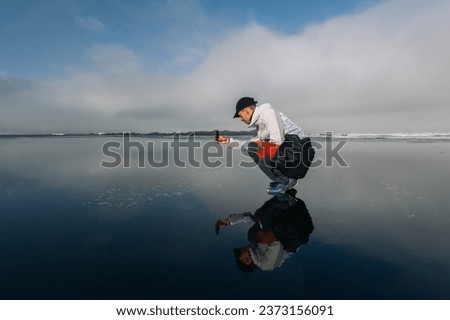 An explorer is crouching on the ocean shore and taking a photo of the horizon. A man is capturing the open sea while crouching on the beach.