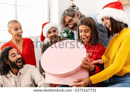 Excited teenage girl opening christmas present with her family at home. Multi generational family celebrating xmas together sitting on sofa in the living room.