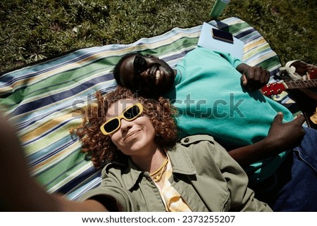 Top view of fun young couple taking selfie outdoors lying on picnic blanket and wearing sunglasses in sunlight, copy space
