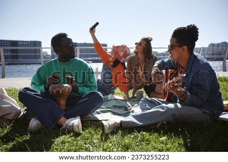 Multiethnic group of young people enjoying picnic on grass in city and playing guitar, copy space