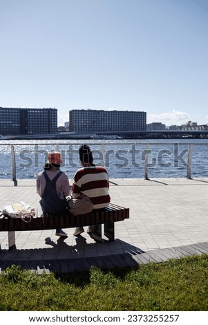 Back view at two young people sitting on bench in riverside park by water with blue sky